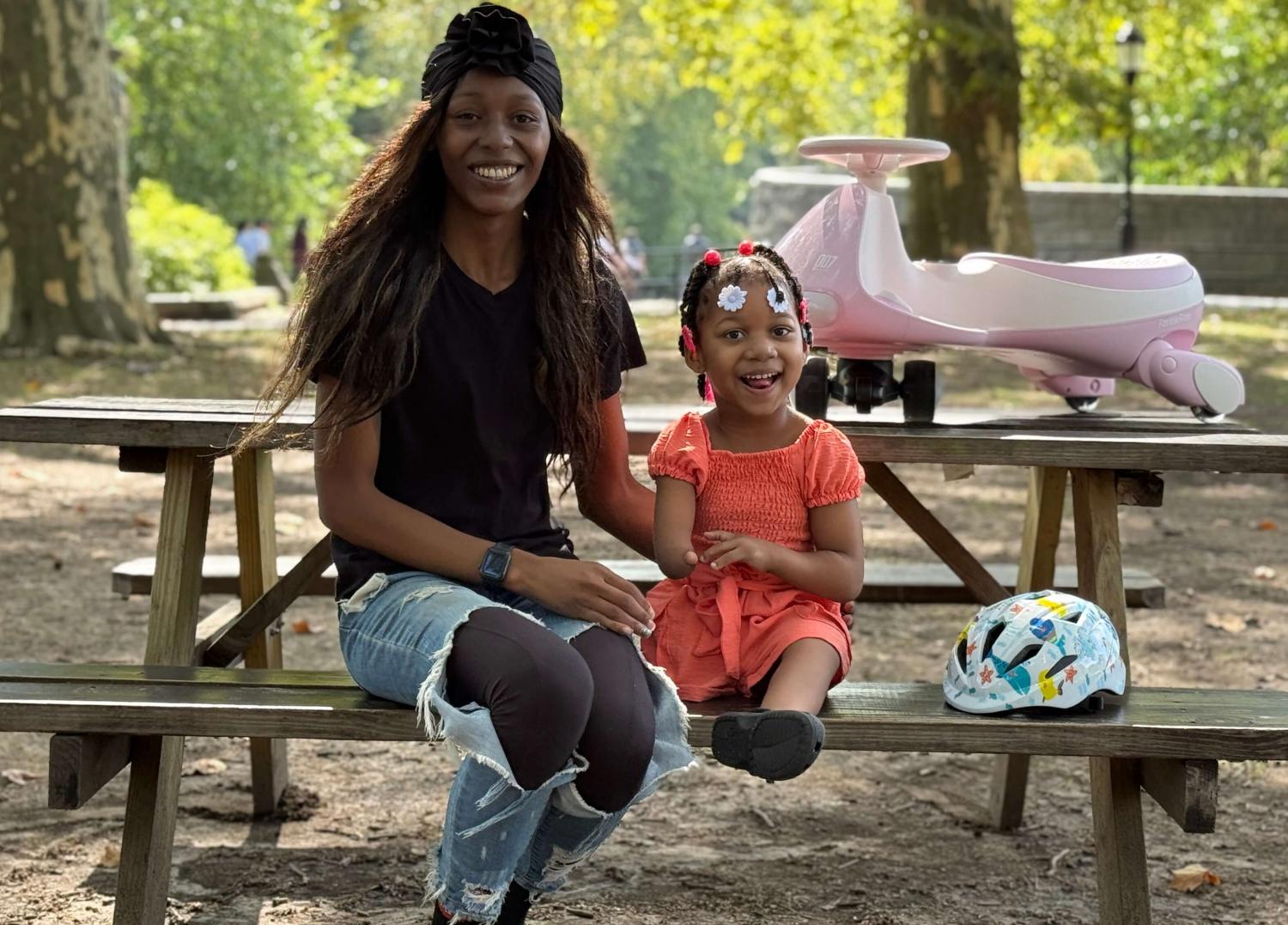 A mother and daughter sit on a park bench, a pink 6KU balance bike on the table. The daughter, Nyla, was born with limb disabilities; she is missing her right hand and right leg.